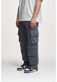 Cargo kalhoty v tmavě šedé tkanině s více kapsami, uvolněný střih a zipové detaily na kotnících, doplněné černobílými teniskami.