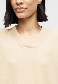 Sweat-shirt beige à col en V fabriqué en tissu doux et texturé. Comprend un détail de couture subtil et une coupe décontractée.