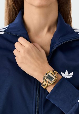 Digitaal horloge met een rechthoekig gezicht, contrast zwart met geel tijgerstreepband, gedragen om de pols. Navy trainjasje met rits en witte accenten.