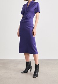 Fialové upnuté šaty s krátkými rukávy, hlubokým V-výstřihem a nařasenými detaily v pase. V kombinaci s černými kotníkovými botami s metalickými akcenty.