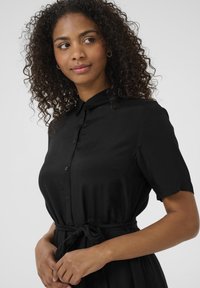 Robe noire en tissu lisse, avec un col, des manches courtes, une fermeture à boutons et une ceinture à nouer à la taille.