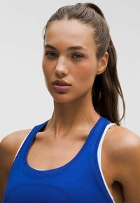 Blauw tanktop gemaakt van gestructureerde stof met een ronde halslijn en witte schouderaccenten. Glad huidtype en lang haar in een paardenstaart gestyled.