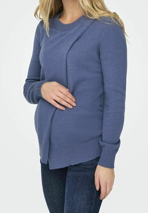 Femme portant un pull bleu à manches longues et un jean foncé, tenant sa main sur son ventre de femme enceinte sur un fond uni.