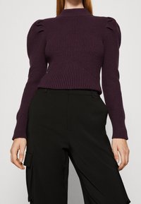 Fialový žebrovaný svetr s vysokým límcem a nafouklými rukávy, ve spojení s černými kalhotami s nákladním kapesem. Texturovaná pletenina.