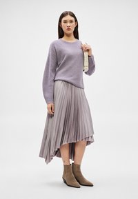 Moteris dėvi alyvinį megztinį, šviesiai violetinę klostuotą asimetrišką sijoną, smėlio spalvos ankletinius batus ir laiko šviesios smėlio spalvos rankinės diržą.