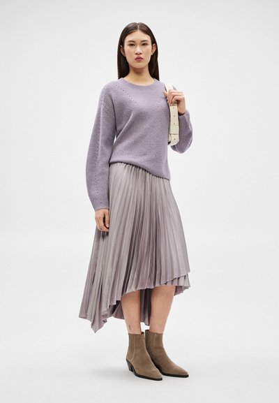 Femme portant un pull en tricot lilas, une jupe plissée asymétrique violet clair, des bottines beige clair, et tenant la anse d'un sac à main beige clair.