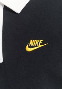 Tejido negro con un cuello blanco que presenta un logotipo de Nike bordado en amarillo y el swoosh en la zona del pecho.
