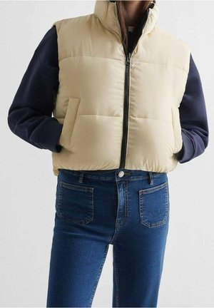 Kvinde iført en beige polstret vest over en marineblå langærmet skjorte, med hænderne i vestens lommer og blå denimjeans.