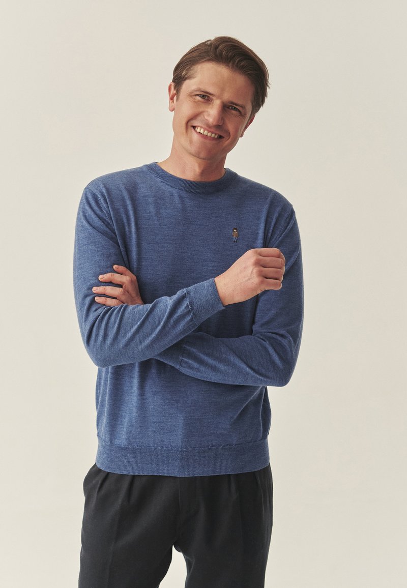 Pull bleu en tricot à manches longues, col rond, avec une petite figure brodée sur la poitrine. Associé à un pantalon noir.