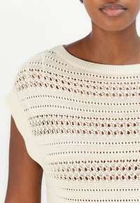 Strikket topp i kremfarge med vid hals og korte ermer, med en åpen tekstur, stripete mønstre og subtile detaljer.