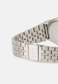 Montre en métal argenté avec un bracelet tressé, comportant un fermoir gravé "CASIO". La montre a un boîtier rond avec un fond texturé.