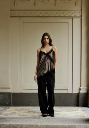 Kobieta stojąca w brązowym satynowym topie bez rękawów z czarną koronkową lamówką oraz luźnych czarnych spodniach na tle dekoracyjnej beżowej ściany.