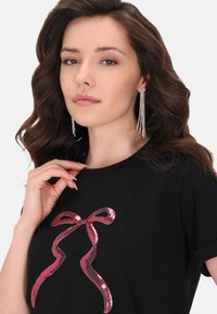 Sort t-shirt med et metallic pink bue-design. Stoffet er glat; modellen bærer lange, glitterende øreringe og har bølget hår.