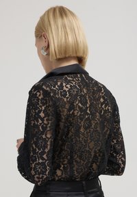 Blonde Person mit kurzen Haaren, die große silberne Creolen und ein durchsichtiges schwarzes florales Spitzenoberteil mit satiniertem Kragen und Manschetten trägt.
