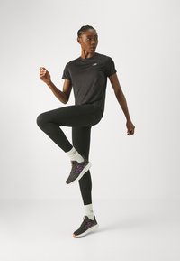 T-shirt de sport noir, leggings noirs et baskets noires avec des accents violets. Modèle posé avec une jambe levée, portant des chaussettes blanches.