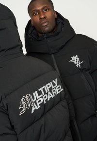 Giacca puffer nera con ampie sezioni trapuntate, colletto alto e logo "MULTIPLY APPAREL" in materiale riflettente.