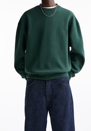 Persona con sudadera oversize verde oscuro, camiseta blanca, collar de cadena plateada y vaqueros sueltos de mezclilla azul oscuro sobre fondo blanco.