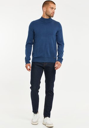 Mies, jolla on päällään sininen pooloneule, tummat farkut ja valkoiset tennarit, seisoo ja katsoo sivulle valkoisella taustalla.