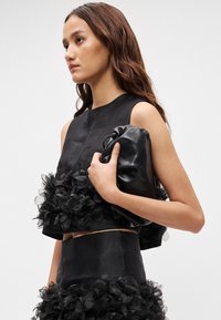 Top preto cropped com detalhes em tule floral, emparelhado com uma saia preta de cintura alta. O modelo segura uma mala de mão preta em couro. Textura suave.