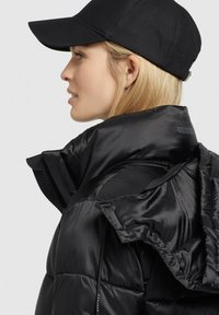 Sort dunjakke med en blank finish, høj krave og lynlås-lomme. Kombineret med en sort cap, der viser et strømlinet, moderne design.