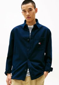 Camicia blu navy con bottoni, dotata di tasca e patch del logo, indossata sopra una camicia a righe, con maniche arrotolate e una vestibilità rilassata.