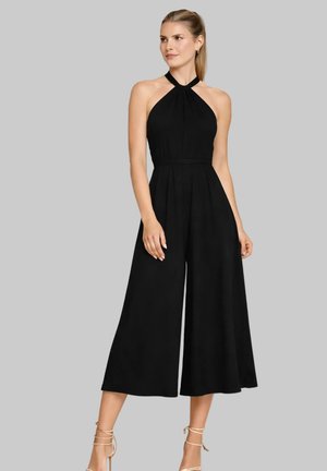 Donna che indossa una tuta nera senza maniche con collo all'americana e pantaloni larghi a gamba corta, con tacchi beige con lacci alla caviglia, in piedi su uno sfondo grigio.