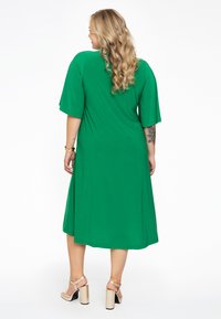 Groene, knielange jurk met korte mouwen, gemaakt van lichtgewicht stof. Heeft een relaxte pasvorm en ronde halslijn. Gepaard met gouden block-heeled sandalen.