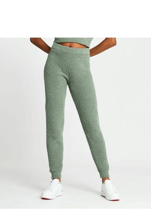 Mujer con pantalones jogger texturizados verdes y zapatillas blancas, de pie con las manos detrás de la espalda contra un fondo liso.