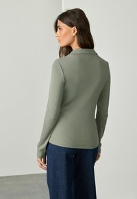 Woman facing sideways wearing a fitted long-sleeve olive green top with a collar and dark blue wide-leg pants against a plain background.