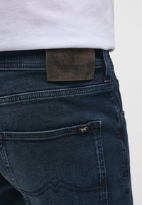 Jeans de mezclilla azul oscuro con un parche de cuero etiquetado como "MUSTANG" en la cintura; presenta detalles de costura y una pequeña etiqueta de logo en el bolsillo.