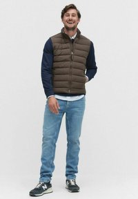 Brązowa kamizelka puchowa na ciemnogranatowej koszuli z długim rękawem, jasnoniebieskie dżinsy i czarne sneakersy. Posiada zamek błyskawiczny oraz boczne kieszenie.