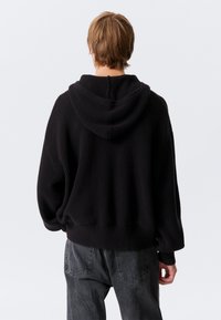 Fekete kötött kapucnis pulóver mintás felülettel, bő szabással és tágas kapucnival. Bordázott szegély és mandzsetta jellemzi, sötétszürke nadrággal párosítva.