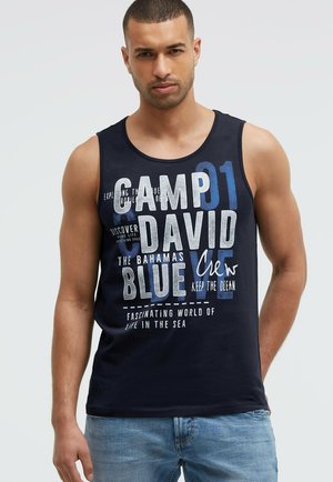 Man wearing a navy blue sleeveless tank top with white and blue text, paired with light blue jeans, standing against a plain background.