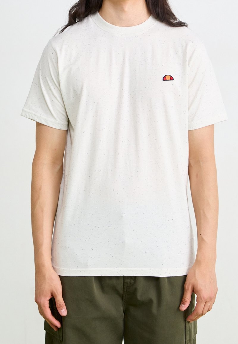 Personne portant un t-shirt blanc à manches courtes tacheté avec un petit logo rouge et orange sur la poitrine et un pantalon vert olive.