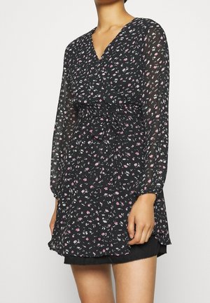 Vrouw draagt een zwarte lange mouwen bloemen wikkeljurk met kleine roze en witte bloemen, gestrikt in de taille over een zwarte rok.