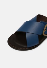 Sandale en cuir bleu avec design à brides croisées, semelle marron et coutures marron contrastantes. Dotée d'une boucle en métal pour ajustement.