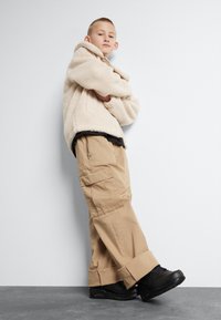 Krém színű bolyhos kapucnis pulóver magas gallérral, bézs színű cargo nadrággal, amely zsebekkel rendelkezik, és fekete, mintás talpú sportcipővel párosítva.