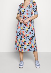 Florales Midi-Kleid in Hellblau mit V-Ausschnitt, kurzen Ärmeln und einem ausgestellten Rock, der mit gelben, roten und schwarzen Blattmustern verziert ist. Kombiniert mit hellblauen Sneakers.