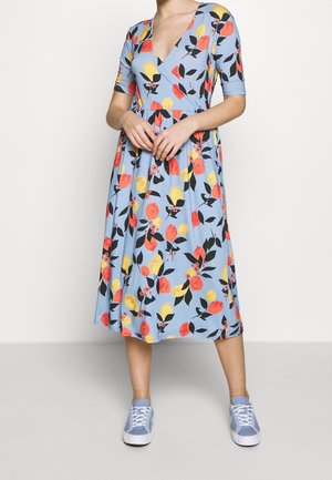Florales Midi-Kleid in Hellblau mit V-Ausschnitt, kurzen Ärmeln und einem ausgestellten Rock, der mit gelben, roten und schwarzen Blattmustern verziert ist. Kombiniert mit hellblauen Sneakers.