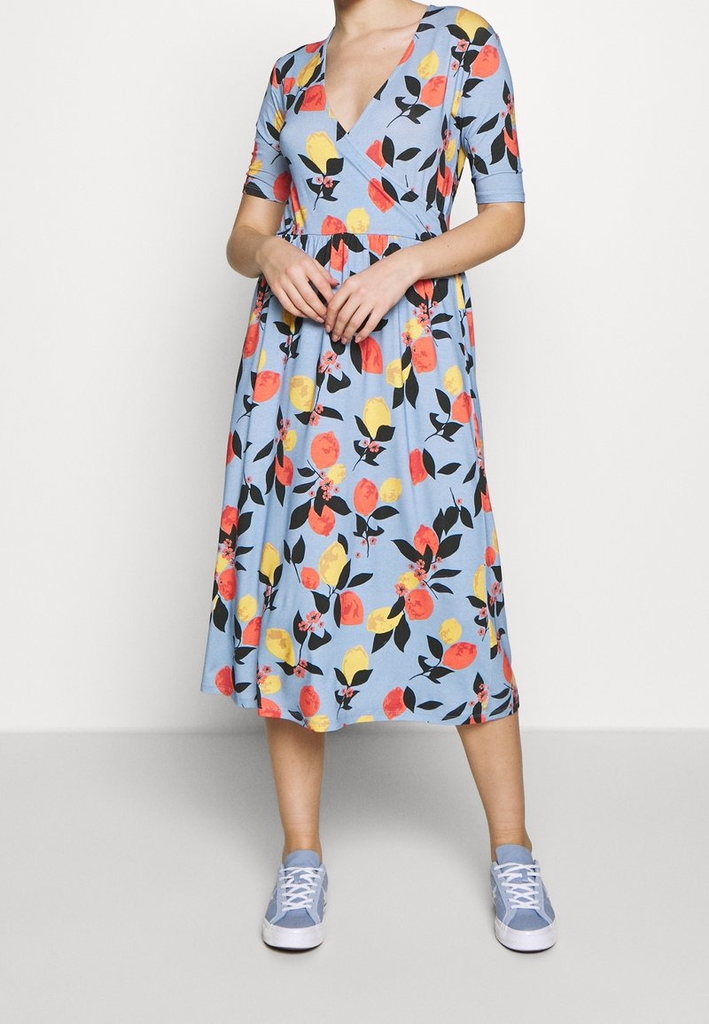 Florales Midi-Kleid in Hellblau mit V-Ausschnitt, kurzen Ärmeln und einem ausgestellten Rock, der mit gelben, roten und schwarzen Blattmustern verziert ist. Kombiniert mit hellblauen Sneakers.