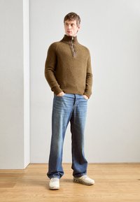 Maglione marrone a maglia con colletto alto e zip, abbinato a jeans azzurri e sneakers bianche, in piedi su un pavimento di legno.