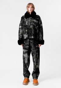 Chaqueta texturizada en negro y gris con un cuello de piel sintética, combinada con pantalones a juego. Cuenta con bolsillos y botas naranjas con una suela gruesa.