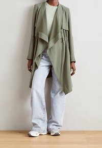 Leichte Jacke in Olivgrün mit drapiertem Vorderteil, langen Ärmeln und Kordelzug-Detail, kombiniert mit hellblauen Jeans in Trompetenform und grauen Sneakern.