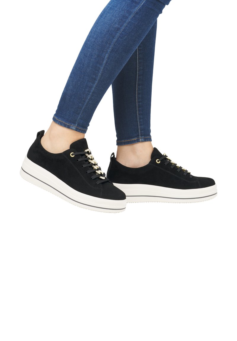 Baskets en daim noir avec une épaisse semelle blanche, dotées d'œillets et de lacets en ton doré, présentées sur une personne portant un jean bleu.