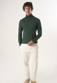 Grüner Rollkragenpullover aus Strickstoff, kombiniert mit weißen Hosen. Einfaches, figurbetontes Design mit langen Ärmeln und glatter Textur.