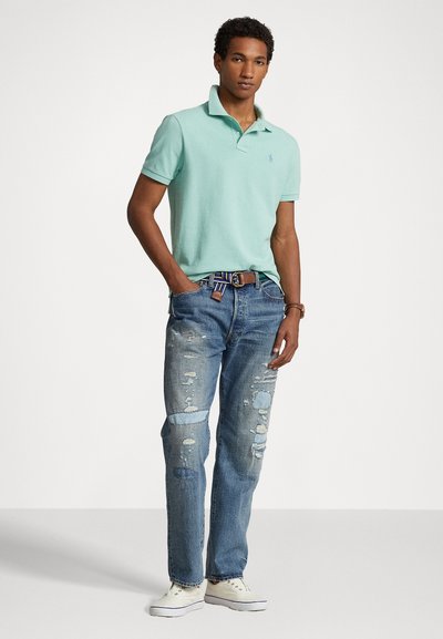 Polo majica v mint zeleni barvi, postarane modre kavbojke z več raztrgami in bele teniske. Rjav pas z večbarvnimi črtami. Model stoji.