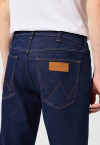 Donkerblauwe denim jeans met gele stiksels, voorzien van een leren Wrangler-logo patch en een opvallend ontwerp op de achterzakken.