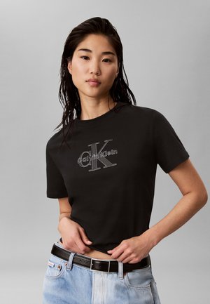 Jeune femme aux cheveux mouillés portant un t-shirt noir Calvin Klein et un jean bleu clair, ajustant son t-shirt à la taille.