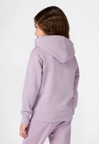 Chica con cabello rizado castaño, vistiendo una sudadera color lila pastel y pantalones a juego, de perfil contra un fondo blanco.