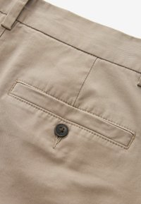 Pantalons en coton beige avec une poche arrière dotée d'un bouton noir unique, de fines coutures le long des bords et d'une texture lisse.